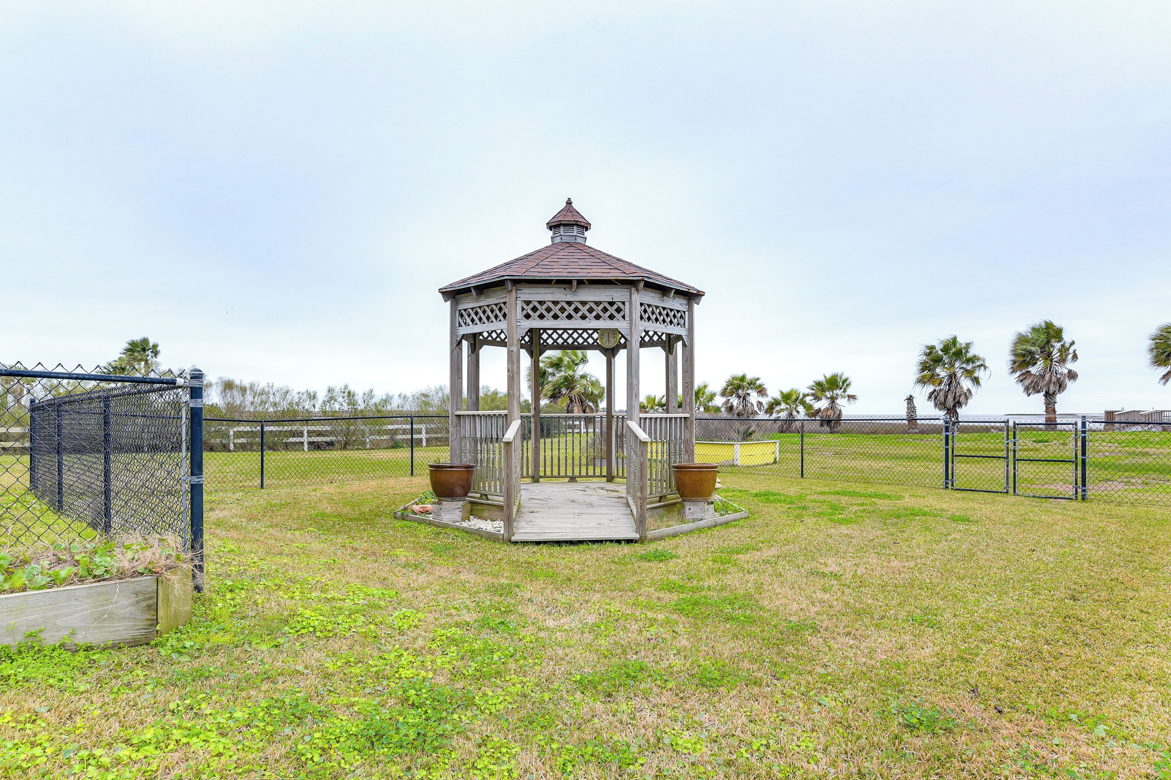 A gazebo with a wooden lattice roof and a central open space, surrounded by a grassy area and palm trees.