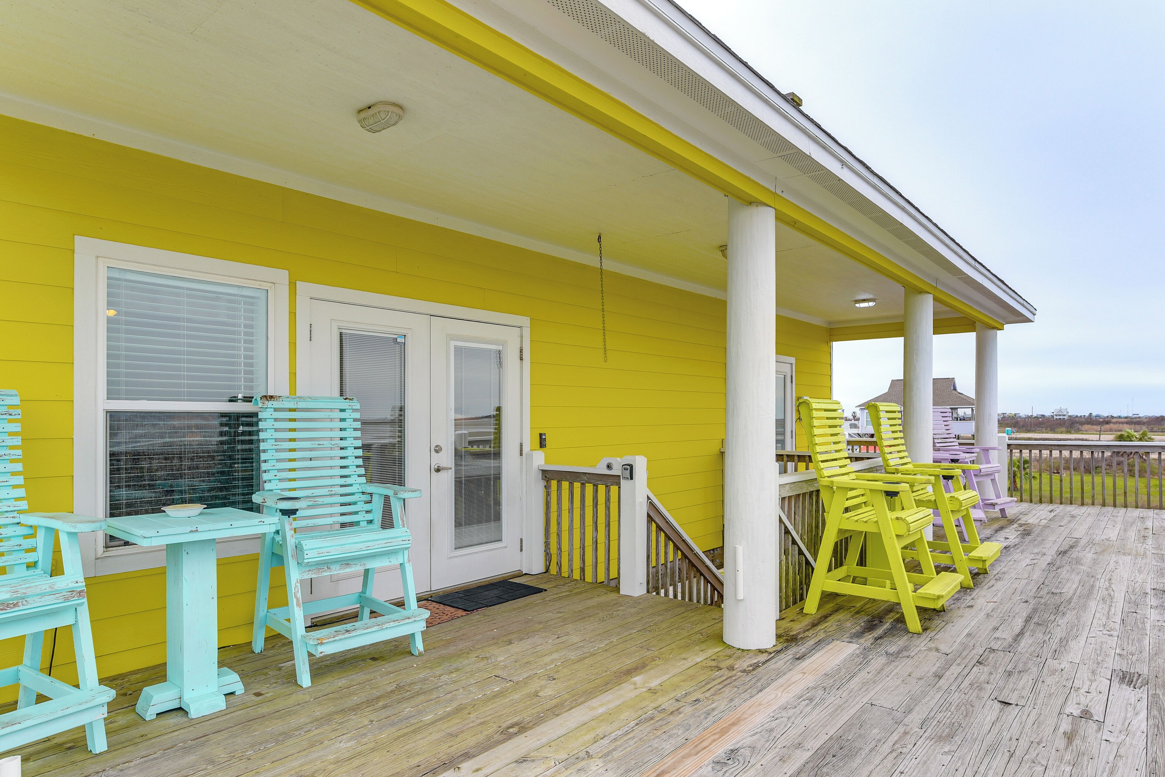 A yellow building with a porch, featuring teal and yellow chairs, a white door, and a wooden deck.