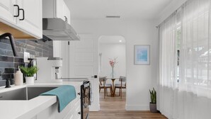 Una cuina moderna amb armaris blancs, una campana extractora d'acer inoxidable i un sòl de fusta que condueix a una zona de menjador amb taula i cadires.