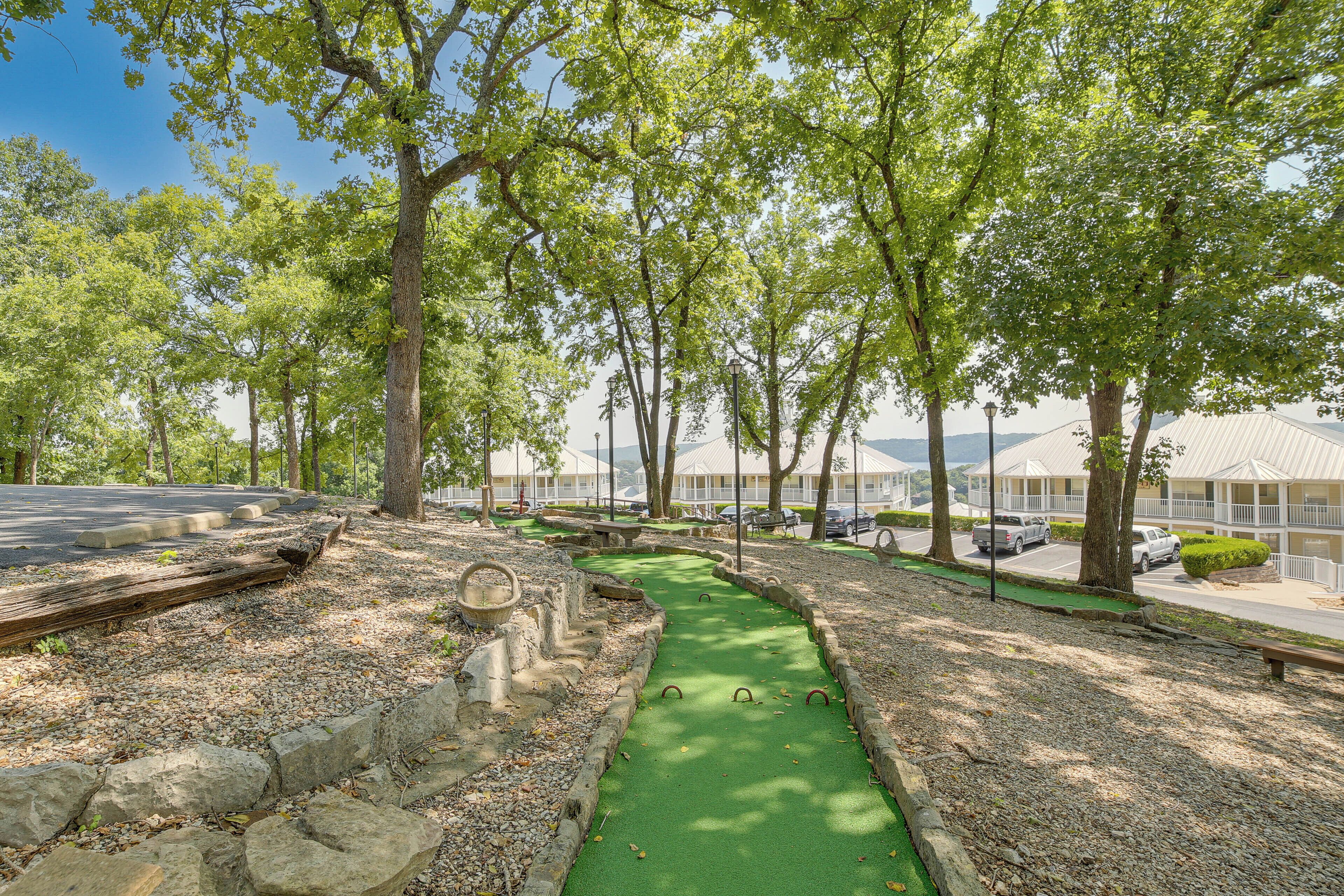 A golf course with a green fairway, trees, and a parking area with buildings in the background.