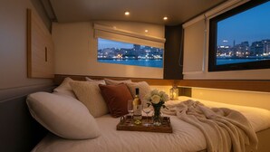 A bed with a city view, a tray with wine glasses and flowers, and a window with a view of the city lights. - Exclusive Boat in Vigo Marina | Private Terrace over the Sea (Vigo)