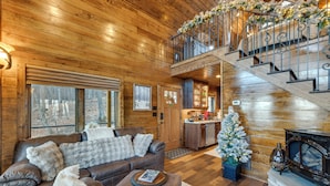 Interior acogedor de una cabaña con detalles de madera, chimenea, escalera y pequeña cocina.