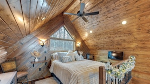 Un dormitorio acogedor en el ático con techo de madera, un ventanal grande, una cama con colcha blanca, un televisor y una barandilla decorativa.
