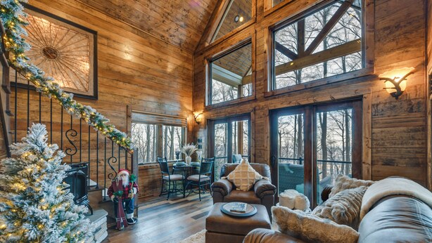 Interior acogedor de una cabaña con un árbol de Navidad, muebles de cuero y grandes ventanales que ofrecen vistas del bosque circundante.