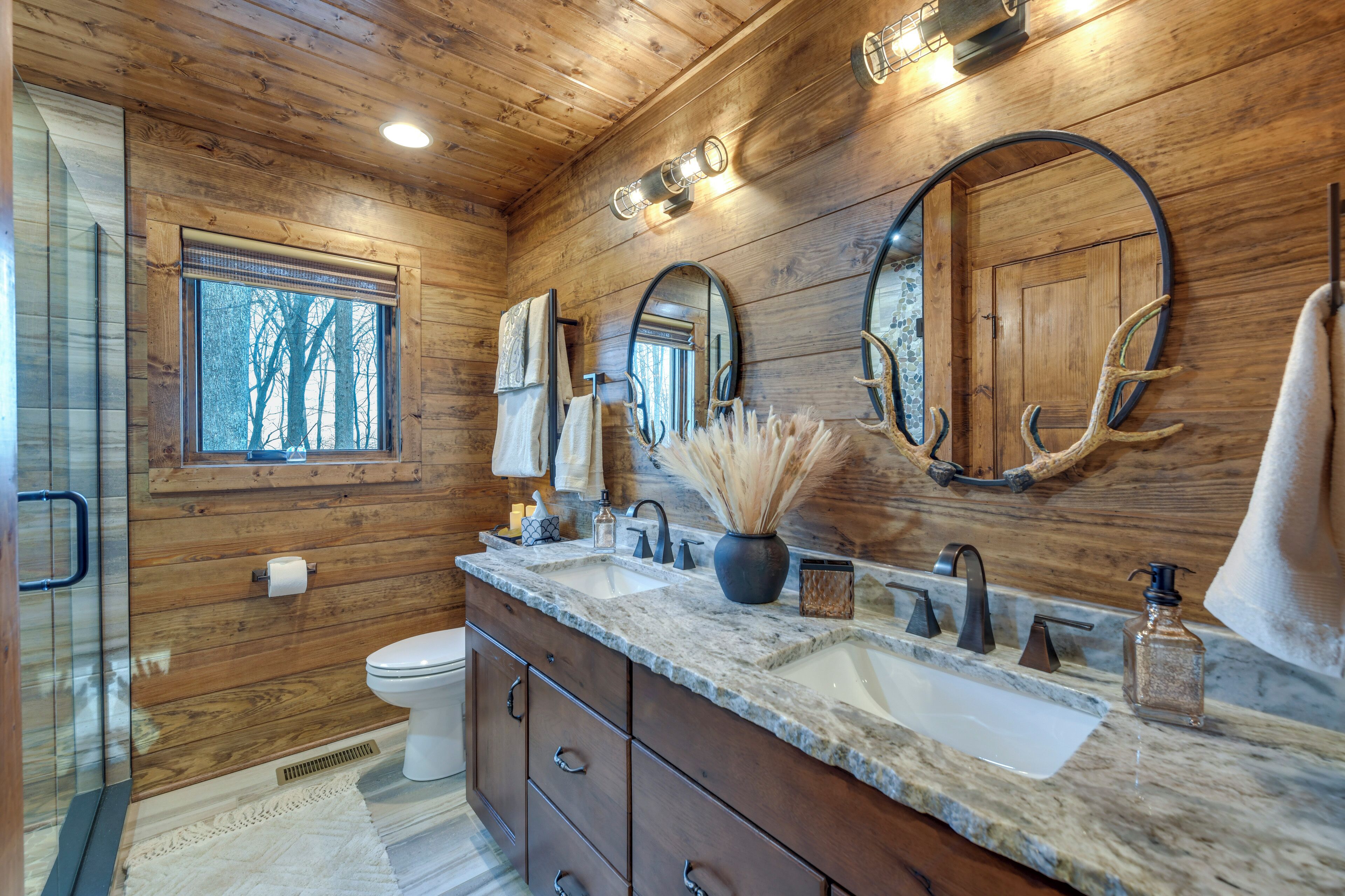 A wooden cabin bathroom with a large window, two sinks, and a mirror.