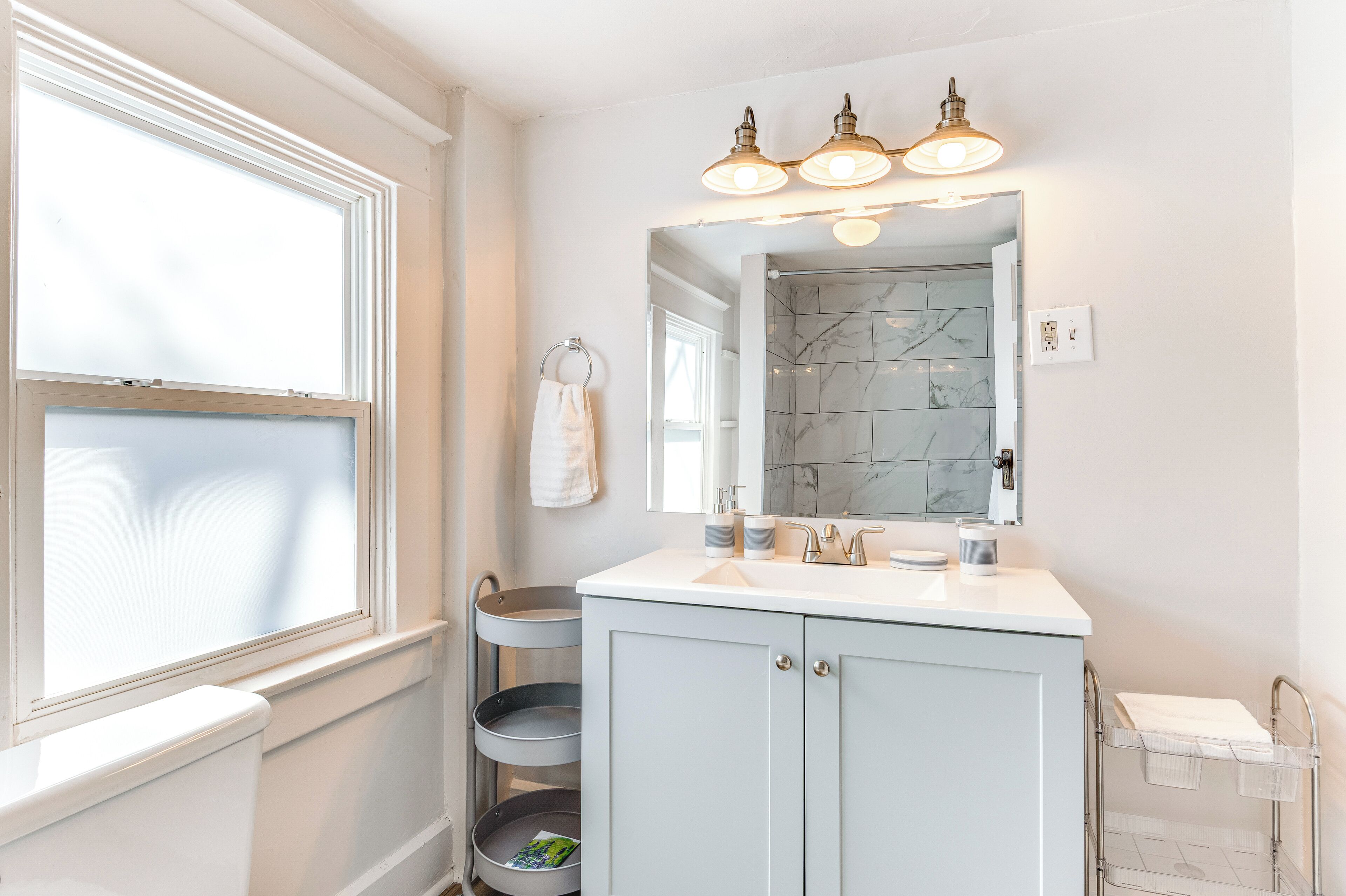 Une salle de bain avec un meuble-lavabo blanc, un miroir et une fenêtre avec des stores.