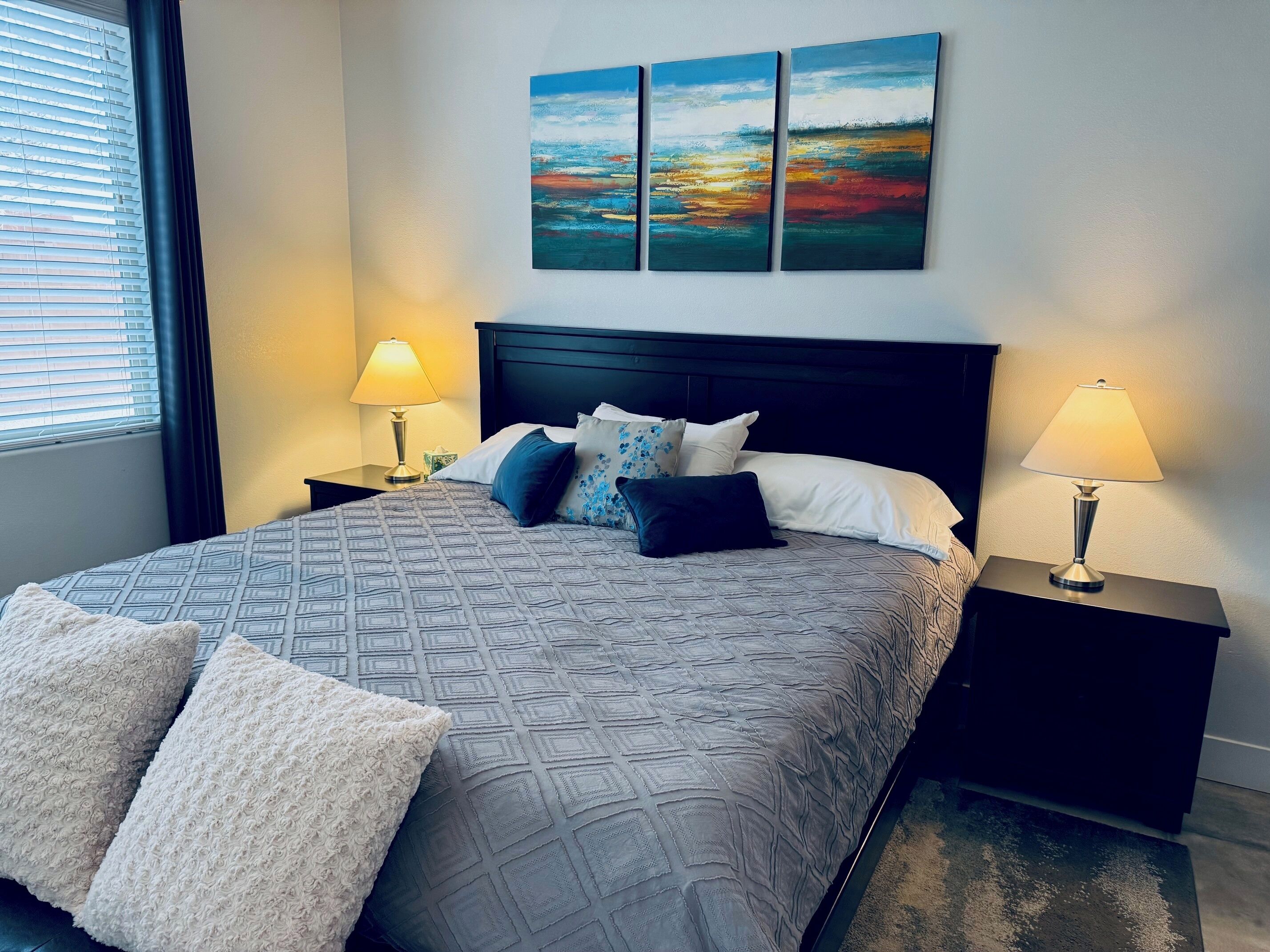 A neatly made bed with a patterned bedspread, two bedside lamps, and a set of three paintings above the bed.