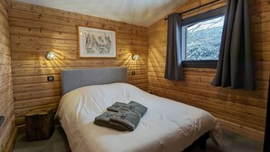 A cozy wooden cabin room with a bed, two wall-mounted lights, a framed picture, and a window with curtains. - Renovated chalet with sauna, near Gérardmer (Sapois)