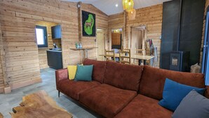 A cozy cabin interior with a wooden log wall, a brown sofa with cushions, a dining table, and a wood-burning stove. - Renovated chalet with sauna, near Gérardmer (Sapois)