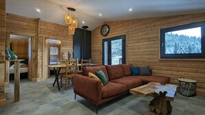 A cozy cabin interior with a wooden log wall, a brown leather sofa, a dining table with chairs, and a large window offering a view of a snowy forest. - Renovated chalet with sauna, near Gérardmer (Sapois)
