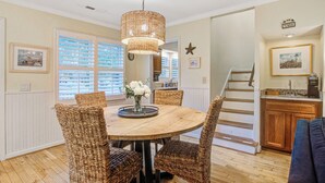 A dining area with a round wooden table, wicker chairs, a hanging light fixture, and a view of the kitchen through a window with blinds.
