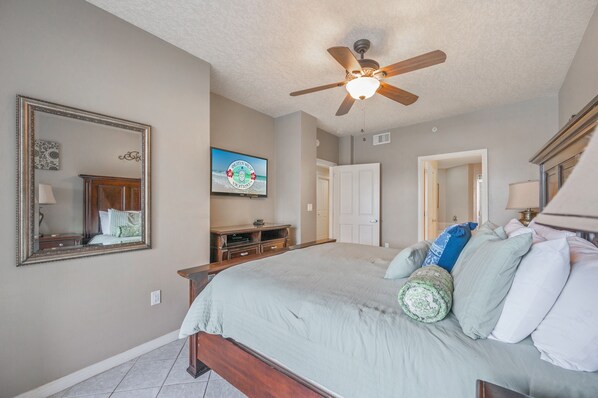 A bedroom with a bed, a ceiling fan, a television, and a door leading to another room. - Gulf 106 Oceanfront 2BR+ bunk room with patio and modern comforts! (Fort Walton Beach)