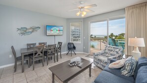 A living room with a dining area, a ceiling fan, a TV, and a view of the ocean. - G316 Gulf-view 2BR+ bunk room w/ balcony, pool access & free beach service (Fort Walton Beach)