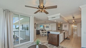 A spacious living area with a large sliding glass door leading to a balcony, a wooden dining table with a vase, and a kitchen with a central island and modern appliances.