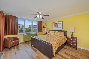 A bedroom with a bed, a nightstand, a dresser, a window with curtains, a ceiling fan, and a view of buildings outside. - Heron 406 | Bay & Lazy River Views | Spacious 3BR Retreat w/ Balcony! (Ft. Walton Beach)