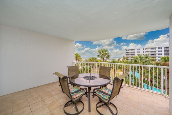 A balcony with a round table and four chairs, overlooking a swimming pool and a building complex. - Heron 406 | Bay & Lazy River Views | Spacious 3BR Retreat w/ Balcony! (Ft. Walton Beach)