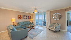 A living room with a sofa, armchair, and coffee table, a ceiling fan, and a large window with curtains. - H602 Top-Floor 3BR Bayfront Condo | Stunning Views | Free Beach Service! (Ft. Walton Beach)