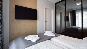 A modern hotel room with a large bed, a flat-screen TV mounted on a geometric-patterned wall, a glass door leading to a bathroom, and a view of the city through the window. - RentPlanet - Perłowa Przystań (Sianożęty)