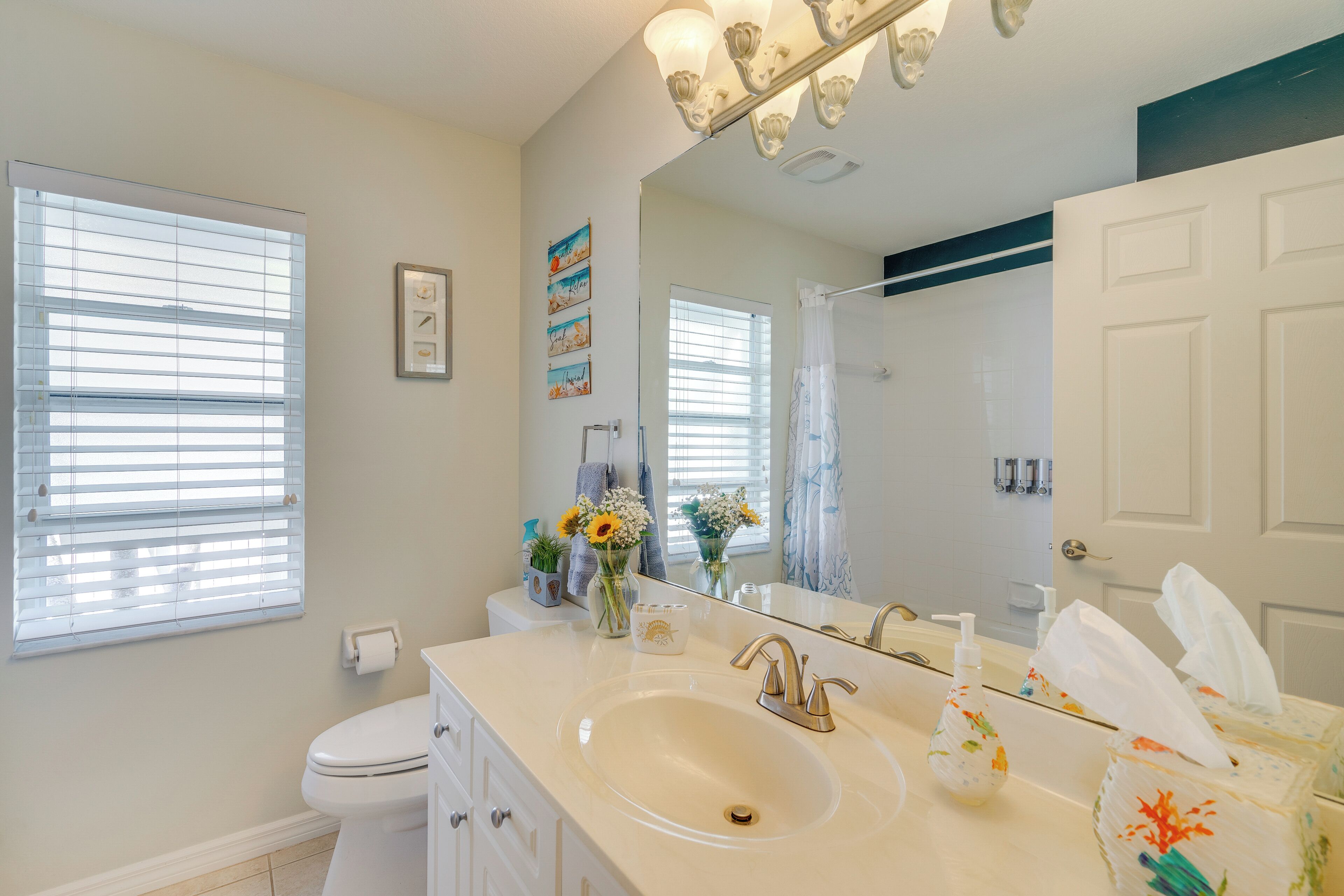 A bathroom with a white sink, vanity, and toilet. There are windows with blinds, a mirror, and a shower curtain.