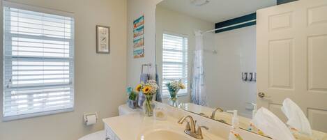 A bathroom with a white sink, vanity, and toilet. There are windows with blinds, a mirror, and a shower curtain.
