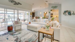 A spacious living room with a large window, a white sofa set, a wooden coffee table, and a decorative rug.