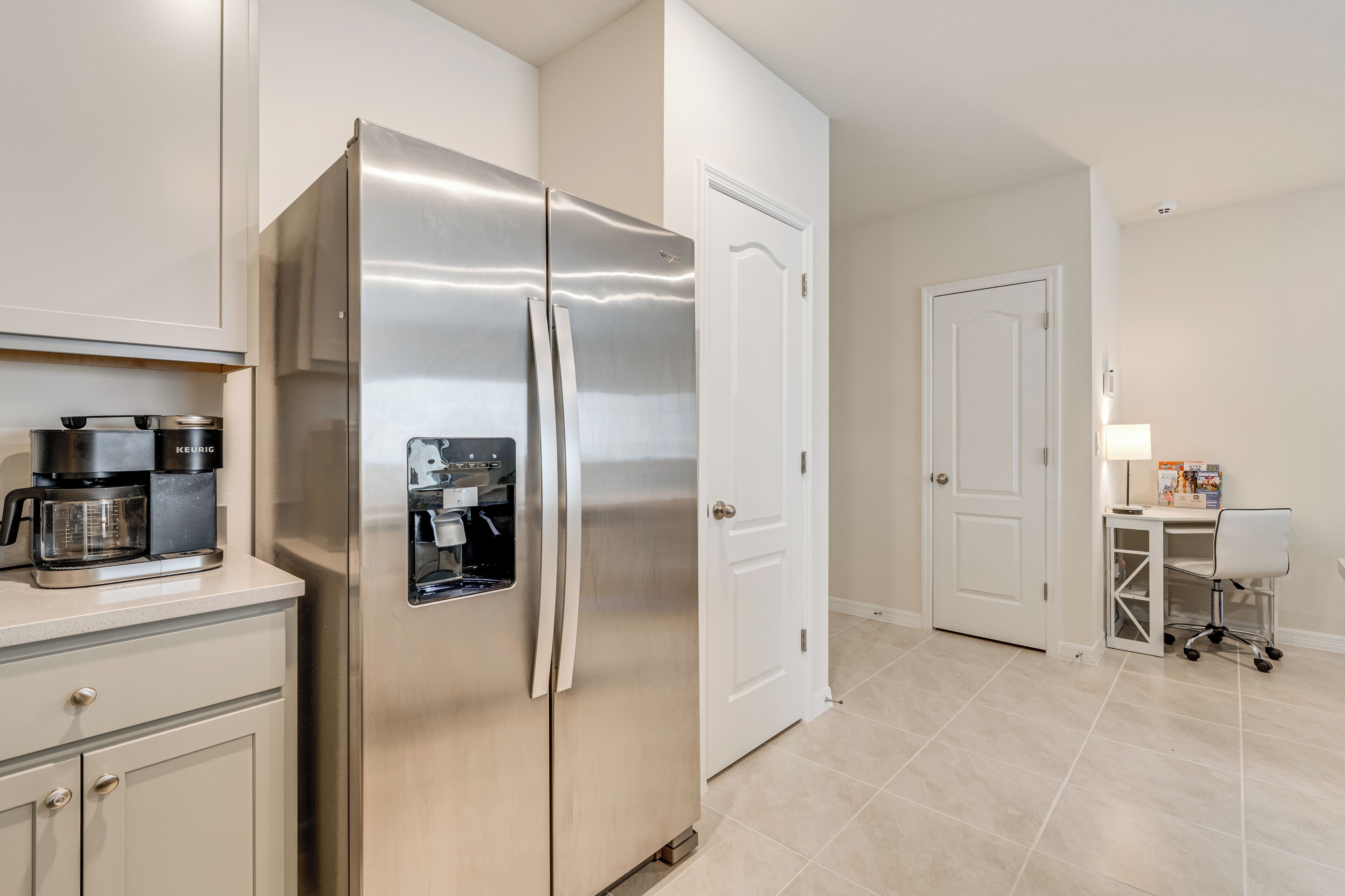 A modern kitchen with a stainless steel refrigerator, a coffee maker, and a small workspace with a chair and desk.