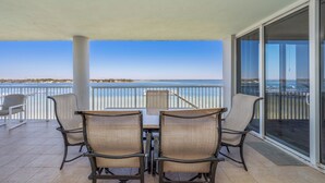A balcony with a table and chairs, overlooking a body of water and a distant shoreline. - O603 Spacious Bayside Condo | Panoramic Views | Lazy River & Resort Access (Fort Walton Beach)