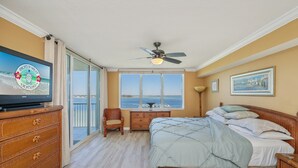A bedroom with a bed, dresser, chair, TV, ceiling fan, and a view of the ocean. - O603 Spacious Bayside Condo | Panoramic Views | Lazy River & Resort Access (Fort Walton Beach)
