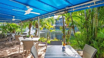 Een terras buiten met tafels en stoelen, een plafondventilator en uitzicht op een waterpartij en groen.