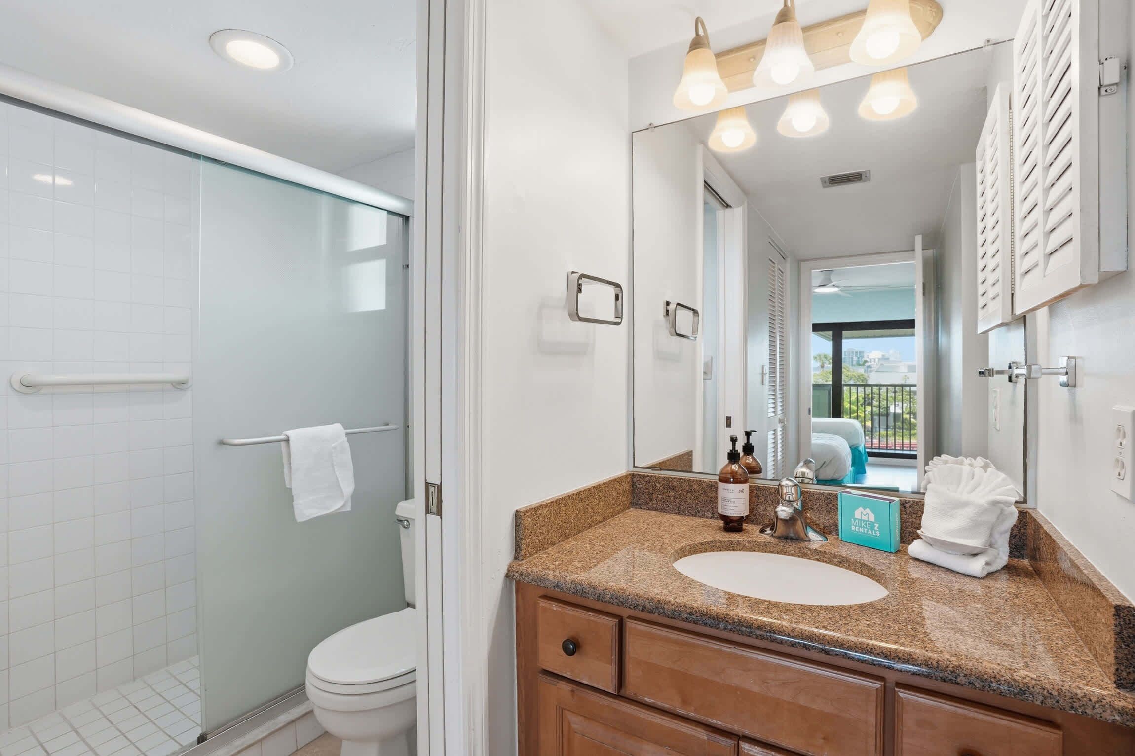 Une salle de bain avec une douche à parois de verre, des toilettes blanches, un lavabo surmonté d’un plan de travail en granit et un grand miroir avec éclairage intégré.