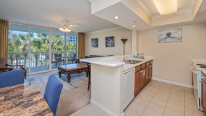 A modern kitchen with a central island, a dining area with blue upholstered chairs, and a large sliding glass door leading to a balcony with palm trees. - Pelican 304 | 1BR Condo w/ Bunks | Lazy River Views & Resort Amenities (Fort Walton Beach)