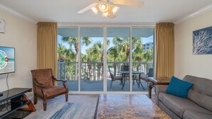 A spacious living area with a ceiling fan, a large sliding glass door leading to a balcony with palm trees, a wooden chair, a sofa, and a flat-screen TV. - Pelican 304 | 1BR Condo w/ Bunks | Lazy River Views & Resort Amenities (Fort Walton Beach)