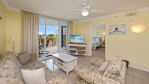 A living room with a sofa, coffee table, and a TV on a stand. There is a sliding glass door leading to a balcony with outdoor furniture.