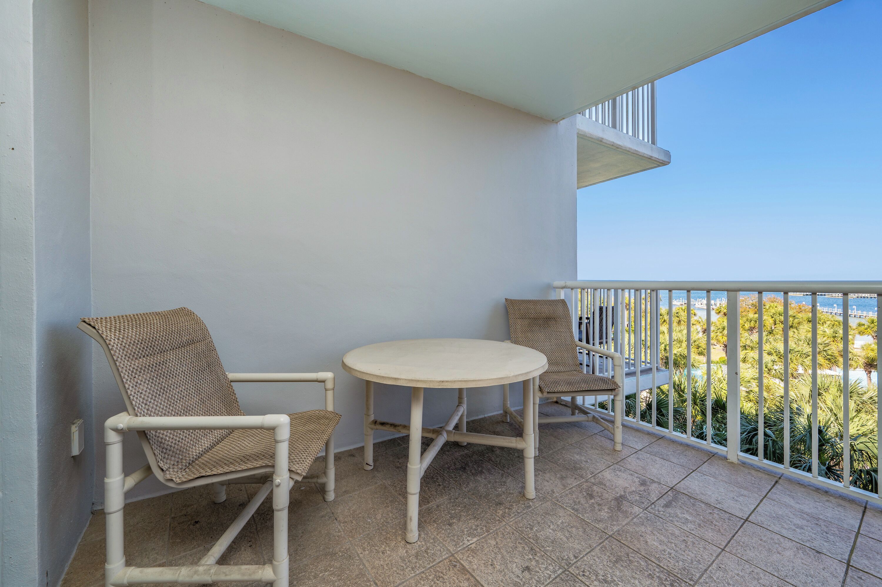 A balcony with two wicker chairs and a small round table, overlooking a view of palm trees and a distant shoreline.