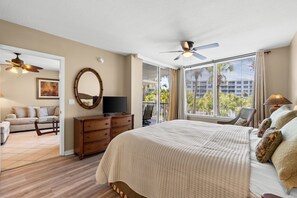 A bedroom with a large bed, a ceiling fan, a TV, a dresser, and a balcony with a view of palm trees.