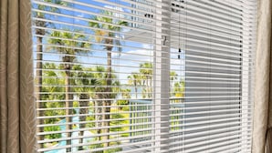 Una ventana con persianas horizontales, vista a palmeras y una piscina.