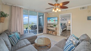 A spacious living room with a large sofa, a coffee table, and a ceiling fan. There is a sliding glass door leading to a balcony with outdoor furniture. - S506 Destin West Sandpiper #506 (Fort Walton Beach)