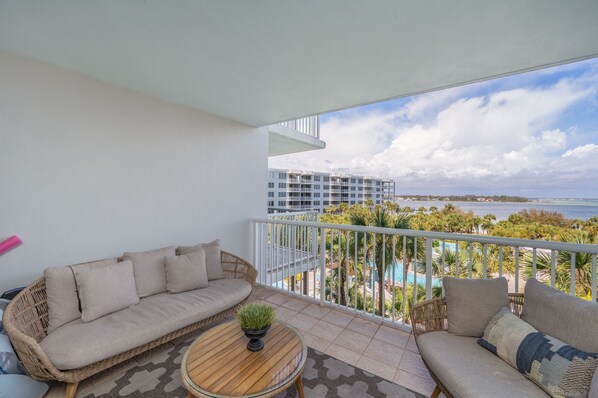A balcony with a wicker sofa and a small table, overlooking a pool and buildings.
