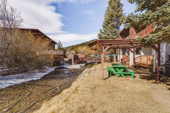 A rustic wooden building with a covered patio and picnic table, surrounded by a natural landscape with a stream and snow patches. - < Half-mi to Red River Ski Area: Townhome w/ Patio (Red River)
