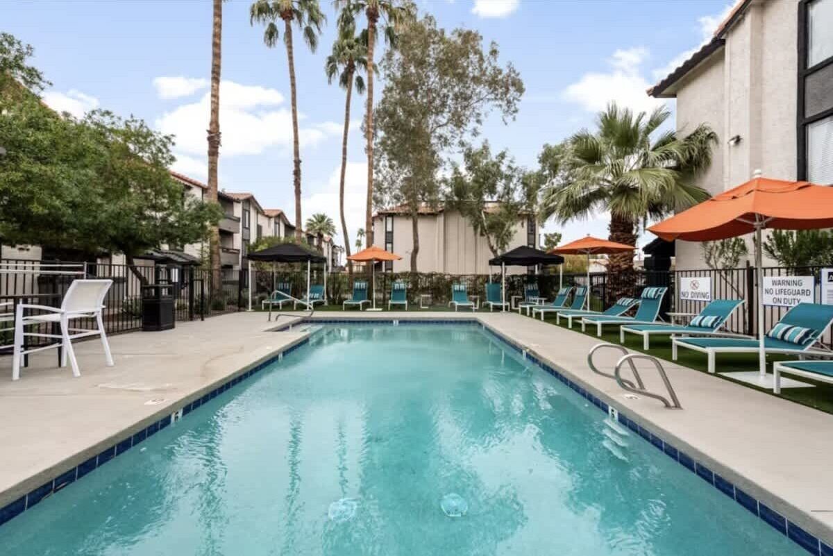 A swimming pool with lounge chairs and umbrellas, surrounded by palm trees and buildings.
