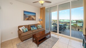 A living room with a brown sofa, a wooden coffee table, a ceiling fan, and a large sliding glass door leading to an outdoor patio with a view of buildings and greenery. - V408 Beachside Villa with Gulf Views, Private Balcony & Resort Pools! (Fort Walton Beach)