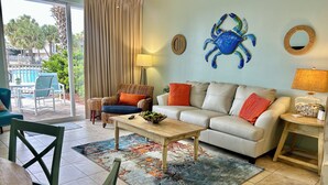 Una sala de estar con un sofá color beige, una mesa de centro de madera y un tapete estampado. Se observa una decoración de pared con forma de cangrejo azul y una lámpara sobre una mesita auxiliar.