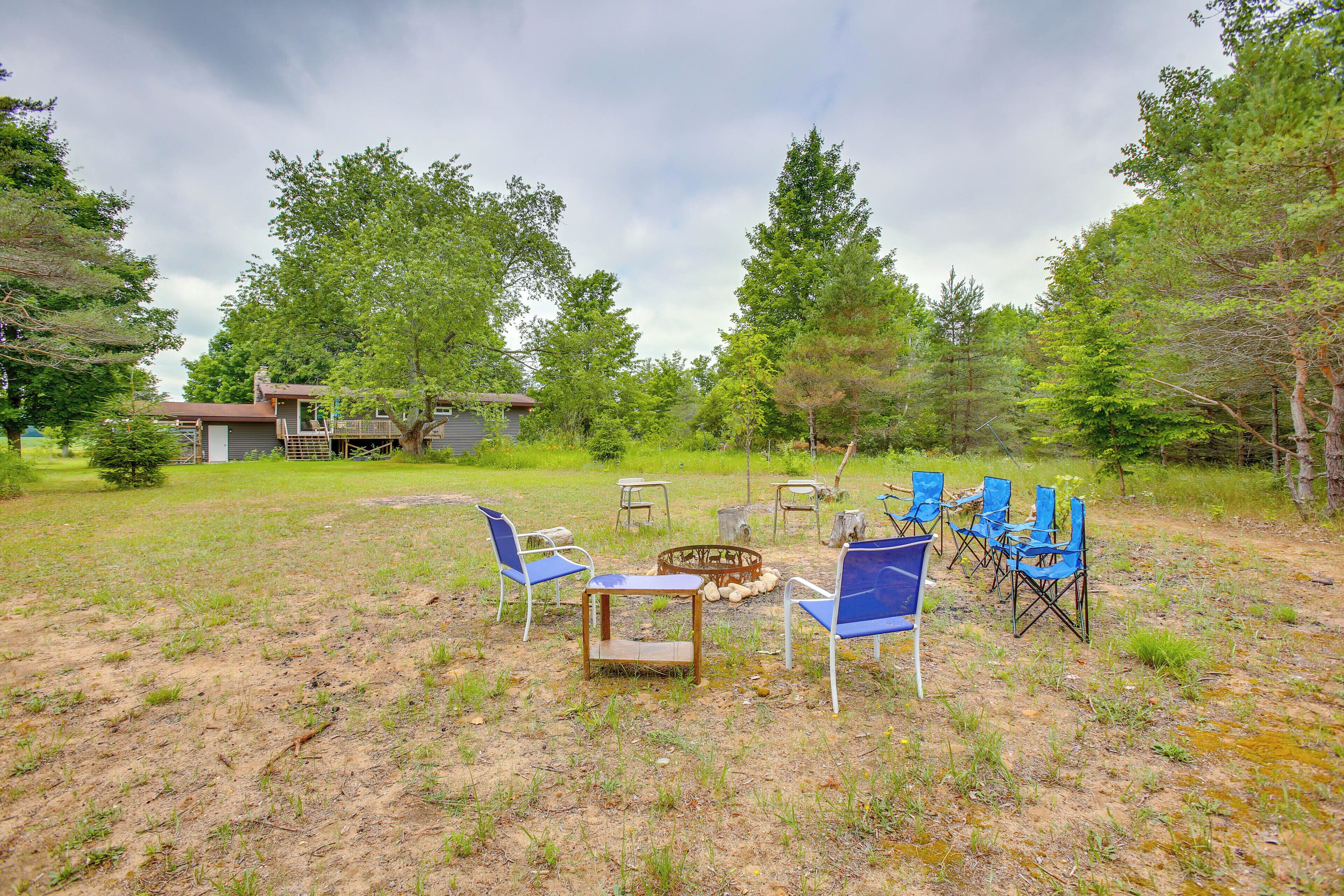A rustic outdoor seating area with a fire pit, surrounded by trees and a house in the background.
