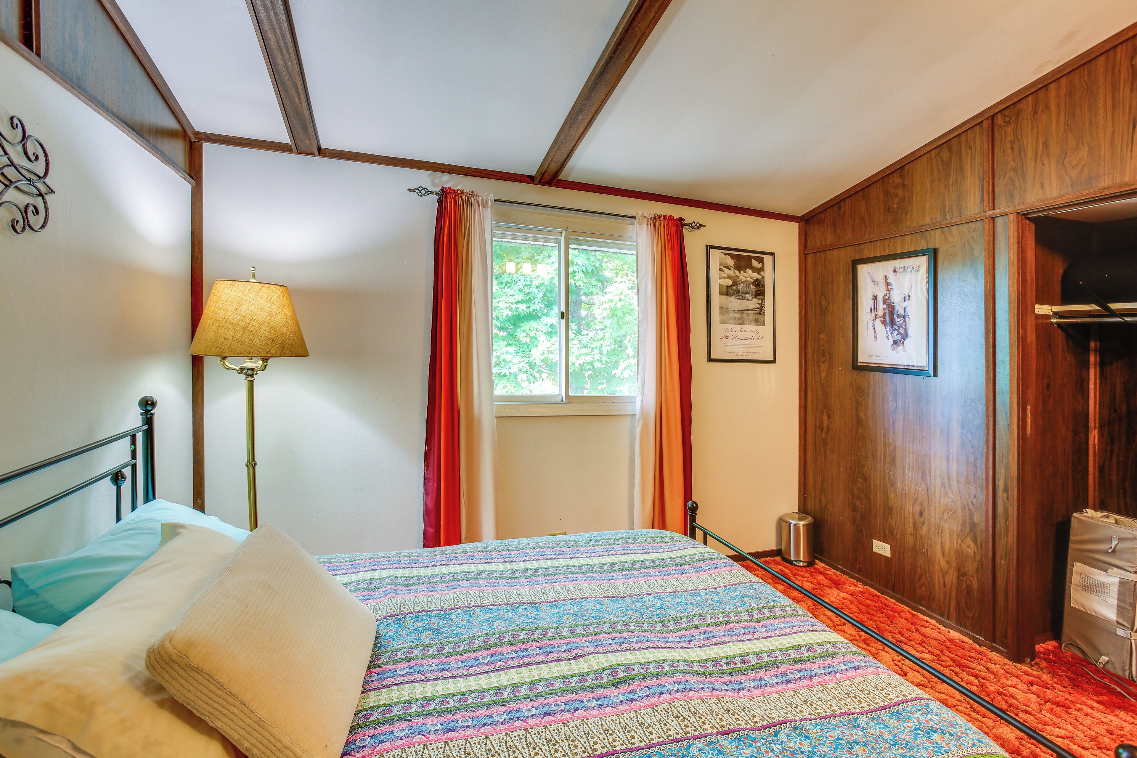 A bedroom with a bed, a lamp, a window with curtains, a framed picture, and a wooden wardrobe.