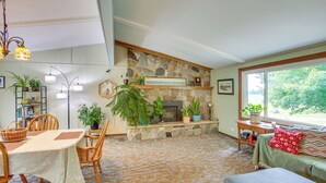 A cozy living room with a stone fireplace, a dining area with a table and chairs, and a large window with a view of greenery. - 8 Mi to Crystal Mountain: Peaceful Up North Home (Copemish)