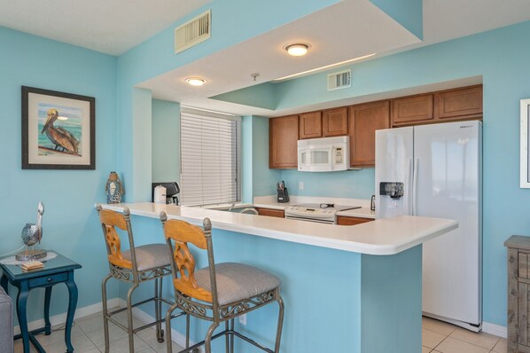 A kitchen with a white island, wooden cabinets, a microwave, and a refrigerator. There are two chairs and a small table with decorative items. - P508 Bayside 1BR Condo | Private Balcony | Lazy River & Beach Access (Fort Walton Beach)