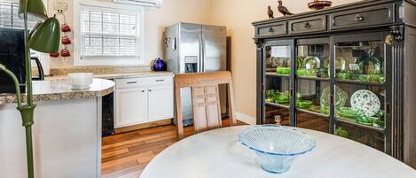 A kitchen with white cabinets, a refrigerator, a dining table with a blue bowl, and a cabinet with glass doors displaying dishes.