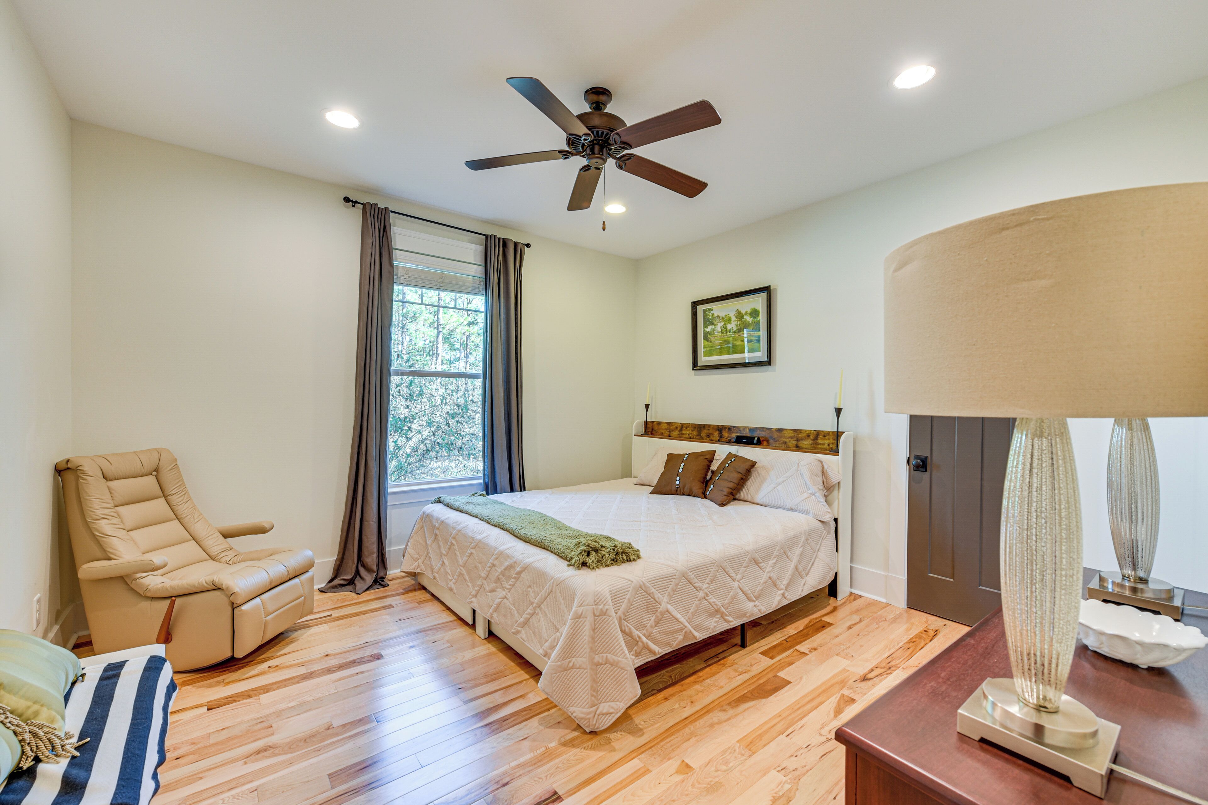 A bedroom with a bed, a reclining chair, a ceiling fan, a window with curtains, and a bedside table with a lamp.