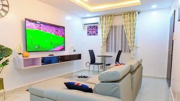 A modern living room with a large flat-screen TV, a sofa, a dining table with chairs, and a window with curtains. - The Mastadon Apartment  (Enugu)