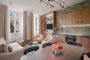 Una sala de estar moderna con un sofá, sillas, una mesa de comedor con fruta y una zona de cocina con armarios de madera y un televisor.